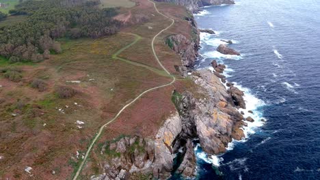 Aerial-drone-shot-of-a-narrow-path-along-steep-cliffs,-in-the-area-of-Morás,-Xove,-Lugo,-Galicia,-Spain-with-sea-waves-crashing-along-the-beach