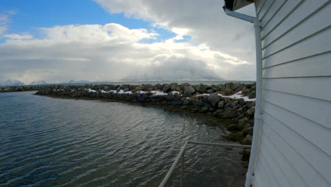 POV-shot-walking-along-the-harbor-building-over-looking-the-breakwater-and-revealing-the-sea,-clouds-and-snow-covered-mountains,-Åse-Harbor,-Andoya,-Norway
