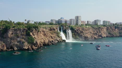 aerial-drone-of-the-waters-of-Lower-Duden-Waterfall-drop-off-a-rocky-cliff-directly-into-the-Mediterranean-Sea-in-Antalya-Turkey-with-large-buildings-overhead-and-local-tourist-boats-viewing-below