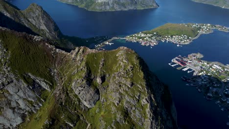 Flying-around-the-peak-of-a-mountain-overlooking-the-little-town-Reine,-the-ocean-and-mountains-in-the-background