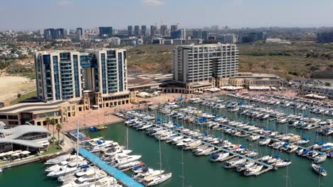 Segelyachten-Fahren-In-Den-Yachthafen-Von-Herzliya-Ein,-Während-Ein-Anderes-Segelboot-Von-Seinem-Liegeplatz-In-Israel-Abfährt,-Während-Mehrere-Touristen-über-Den-Boulevard-Schlendern