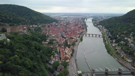 Aerial-dolly-out-of-Heidelberg-village-and-Castle-ruins-next-to-Neckar-River-and-its-bridges-between-hills-covered-in-dense-green-forest,-Germany