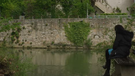 Stylish-woman-on-bench-calmly-watching-duck-passing-by-in-Tubingen-downtown-river-in-Balvaria,-Germany,-Europe