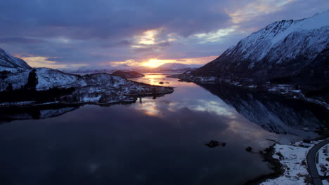 Forward-dolly-aerial-shot-of-sun-setting-on-a-partially-cloudy-day-with-majestic-snow-covered-mountains-and-beautiful-reflection-on-a-calm-waters-of-a-lake-or-sea