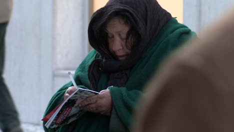 An-old-lady-writes-in-a-magazine,-wearing-a-dark-green-cloak-to-protect-herself-from-the-cold