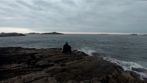 Man-sitting-on-cliff-close-to-ocean-in-moody-light-after-sunset-with-waves-crushing-up-on-shore-around-him---Static-clip-with-thoughtful-person-and-dramatic-cloudy-sky