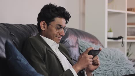 Young-excited-Indian-man-enjoying-playing-games-on-his-smartphone,-couch