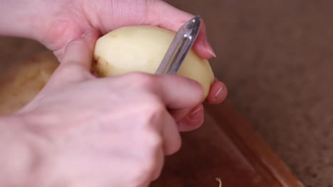 Cerrar-Manos-De-Mujer-Pelando-Patatas-Con-Pelador-De-Verduras-Encima-De-La-Mesa-De-La-Cocina