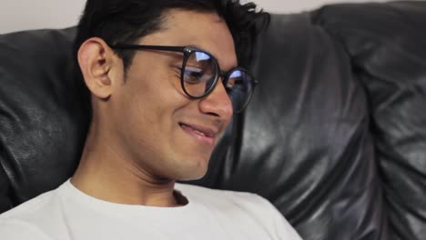 Young-man-with-glasses-sitting-on-couch-and-smiling,-close-up-view