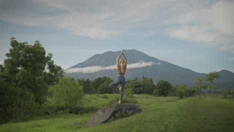 Athletic-blond-woman-in-yoga-tree-pose-balancing-on-rock,-Mount-Agung-in-background