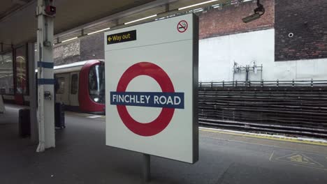 View-Of-Finchley-Road-Roundel-Station-Sign-With-Northbound-Met-Line-Train-Arriving-At-Platform-On-12-May-2022