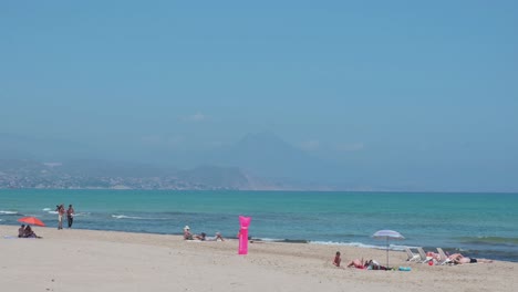 People-sunbathe-while-lying-down-and-have-a-stroll-at-the-beach-on-the-shore-of-the-Mediterranean-sea-in-Alicante,-Spain