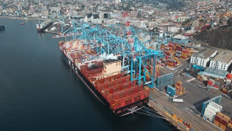 Aerial-orbit-of-a-container-cargo-ship-docked-near-cranes,-vapor-in-air,-ready-to-be-loaded-in-Valparaiso-Sea-Port,-Chile