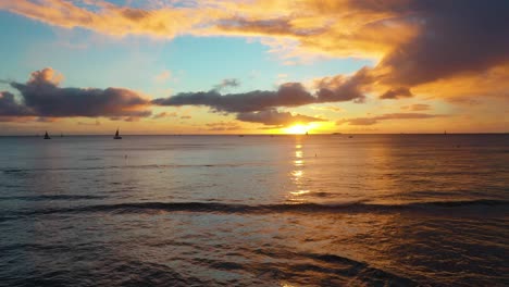Luftdrohne-Fliegt-über-Den-Wunderschönen-Sonnenuntergang-Am-Meer-Mit-Segelbooten-Am-Strand-Von-Waikiki-In-Honolulu,-Hawaii-Zur-Goldenen-Stunde-Mit-Farbenfrohem-Orangefarbenem-Himmel,-Während-Wellen-Gegen-Die-Brechmauer-Krachen