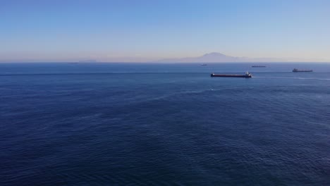 Ein-Großer-Massengutfrachter-Fährt-Von-Algeciras-In-Der-Straße-Von-Gibraltar-Ab-Und-Passiert-Dabei-Einen-Anderen-Frachter-Auf-Dem-Weg-Zum-Atlantik,-Im-Hintergrund-Die-Küste-Afrikas-Und-Vor-Anker-Liegende-Schiffe