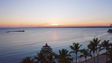 Luftaufnahme,-Die-Das-Ruhige-Karibische-Meer-Und-Die-Silhouette-Von-Palmen-Am-Strand-Und-An-Der-Fußgängerbrücke-Bei-Sonnenuntergang-Zeigt