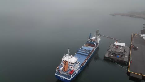 Unloading-Fish-From-Cargo-Ship-On-A-Foggy-Day-In-Faskrudsfjordur,-East-Iceland---aerial-drone-shot