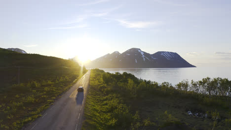 Car-driving-a-small-road-into-the-sunset-next-to-a-ocean-and-blue-sky-with-mountain-landscape-in-the-background