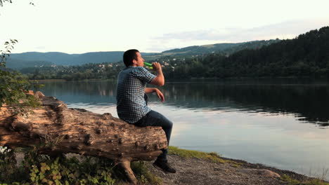 A-man-sits-alone-on-a-tree-stump-looking-out-over-a-lake-as-he-drinks-a-beer