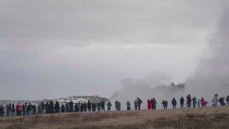 Static,-wide-shot,-of-lots-of-people-watching-the-erupting-geisir,-on-a-cloudy,-rainy,-autumn-day,-in-Geysir,-Iceland
