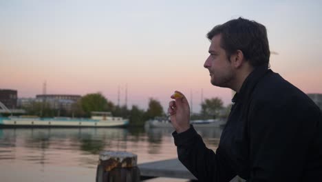 Wide-shot-of-a-young-man-snacking-on-an-orange-slice-and-smiling-on-a-water-pier-during-sunset