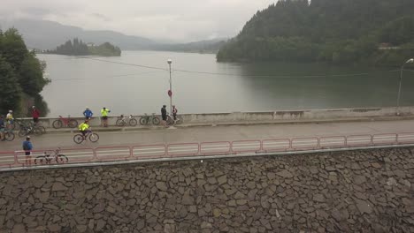 Aerial-view,-orbit-style,-of-a-group-of-cyclists-standing-over-the-dam-at-Colibita-Lake-while-one-cyclist-peddles-along,-during-Tura-Cu-Copaci-race