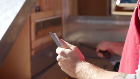 Close-up-shot-of-the-hands-of-a-construction-worker-using-a-large-rough-metal-file-to-grind-and-smooth-a-piece-of-sheet-metal-angle-iron-then-placing-it-onto-the-tiny-house-travel-trailer
