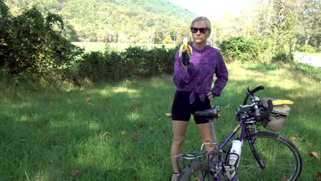 Pretty,-blonde,-mature-woman-with-sunglasses-and-gloves-next-to-her-bicycle-eating-a-banana-on-the-side-of-the-road