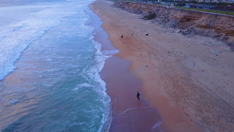 Drohnenaufnahmen-Eines-Surfers,-Der-Bei-Sonnenuntergang-Am-Strand-Spaziert