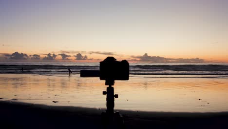 A-camera-is-set-up-on-a-tripod-to-take-pictures-of-the-stunning-view-of-the-sunset-at-low-tide-in-Laguna-Beach-as-surfers-surf-and-the-sun-peaks-out-amongst-rocks-and-reflects-off-the-water