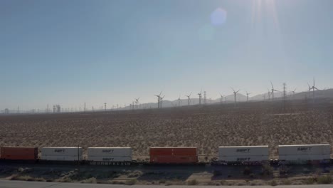 Tren-De-Carga-Moviéndose-Por-Una-Carretera-Del-Desierto-De-Mojave-Con-Turbinas-Eólicas-En-Segundo-Plano