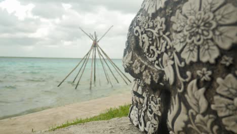 Side-of-mexican-sculpted-skull-with-wooden-tipi-structure-on-lagoon-in-background