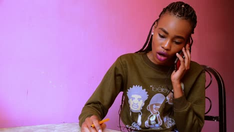 Mujer-Africana-Escribiendo-En-Una-Mesa-Mientras-Contesta-Una-Llamada-Con-Aspecto-Estresado