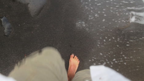 Point-of-view-shot-of-a-man-walking-barefoot-on-a-black-sand-beach-with-waves-hitting-his-walking-feet