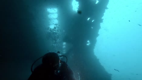 A-scuba-diver-diving-inside-of-an-old-civilian-shipwreck-in-deep-blue-water-,-dark-scenery-lighten-up-with-sunlight-coming-from-the-surface