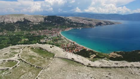 Aerial-Fly-Over-a-mountain-revealing-a-small-coastal-town-of-Baška,-Krk,-Croatia