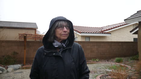 An-elderly-woman-with-glasses-and-a-raincoat-standing-in-a-winter-weather-rain-storm-as-raindrops-fall-in-slow-motion