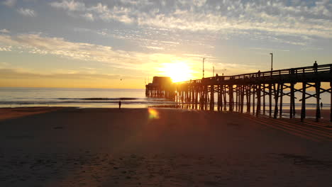 Drohne-Fliegt-Unter-Dem-Newport-Beach-Pier-Bei-Sonnenuntergang-In-Kalifornien-Mit-Sonnenlichtstrahlen-Und-Menschen-In-Silhouette-Auf-Der-Sandantenne