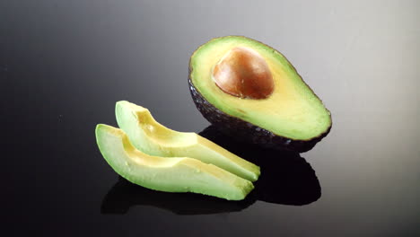 A-ripe-avocado-cut-in-half-with-the-pit-and-two-slices-ready-isolated-on-a-black-mirror-background
