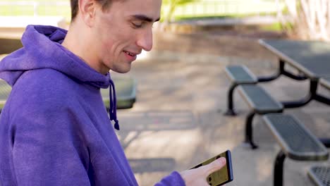 Young-adult-male-browses-social-media-while-chatting-on-phone-in-a-park,-smiling