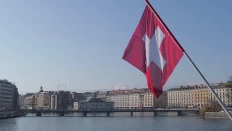 Schweizer-Flagge-Schwebt-Im-Vordergrund-Mit-Genf-Im-Hintergrund-An-Einem-Strahlend-Sonnigen-Tag-Neben-Dem-Fluss