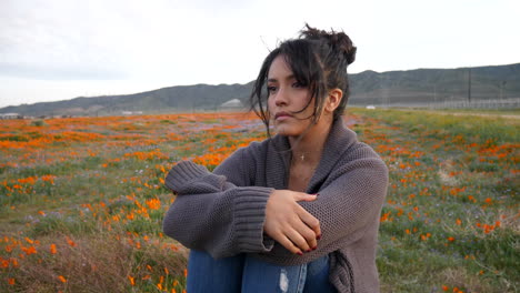 A-cute-and-attractive-girl-looking-sad-and-serious-sitting-alone-in-a-rural-field-of-wild-flowers-with-wind-blowing-her-hair-at-dusk-SLOW-MOTION