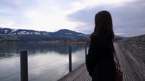Orbiting-shot-around-a-young-asian-woman-sitting-on-a-bench-and-standing-up-to-enjoy-the-beautiful-view-to-the-lake-of-Zurich-in-Rapperswil,-Switzerland