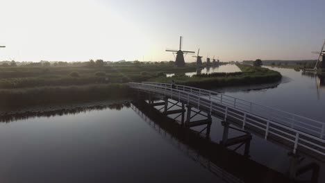 Eine-Drohnenaufnahme,-Die-Nach-Rechts-Schwenkt,-Um-Ein-Modell-Herum,-Das-Auf-Einer-Brücke-Läuft-Und-Bei-Sonnenaufgang-Auf-Holländische-Windmühlen-In-Den-Niederlanden-Blickt