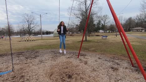 Slow-motion-footage-of-a-beautiful-brunette-college-teenager-swinging-in-a-city-park-wearing-jeans,-a-white-shirt-and-a-black-jacket