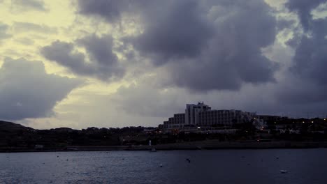 early-morning-in-Qawra,-Malta,-before-sunrise,-rainy-stormy-clouds-over-buildings-and-early-morning-traffic