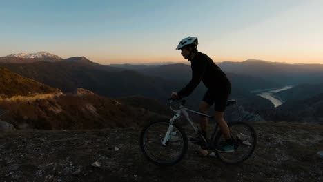 Hombre-Montando-En-Bicicleta-En-La-Cima-De-Una-Montaña-Con-Un-Hermoso-Lago-Del-Cañón-Al-Atardecer-En-El-Fondo