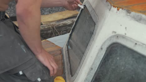 Young-carpenter-using-scraper-to-flatten-sika-flex-mastic-sealant-on-windows-on-bow-of-wooden-boat