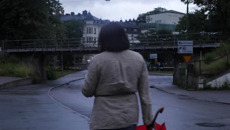 Frau-Mit-Beiger-Jacke,-Die-Mit-Einem-Regenschirm-Und-Ihrem-Telefon-In-Der-Hand-In-Richtung-Der-Regnerischen-Stadt-Borås-Vorbeigeht