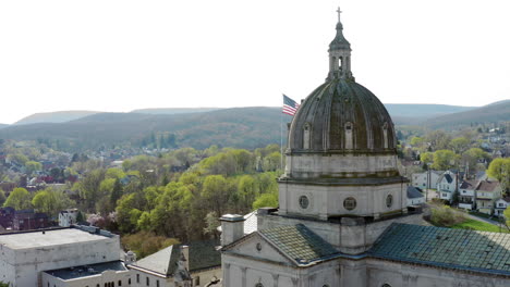 Luftdrohnenansicht-Der-Kathedrale-Des-Allerheiligsten-In-Altoona,-Pennsylvania,-Mit-Der-Amerikanischen-Flagge-Dahinter